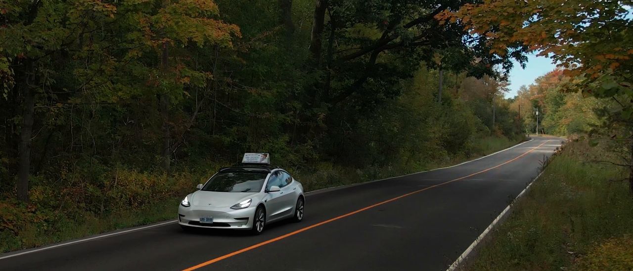 A white GR8 Drivers car with a roof sign, driving a tree-lined road in autumn.